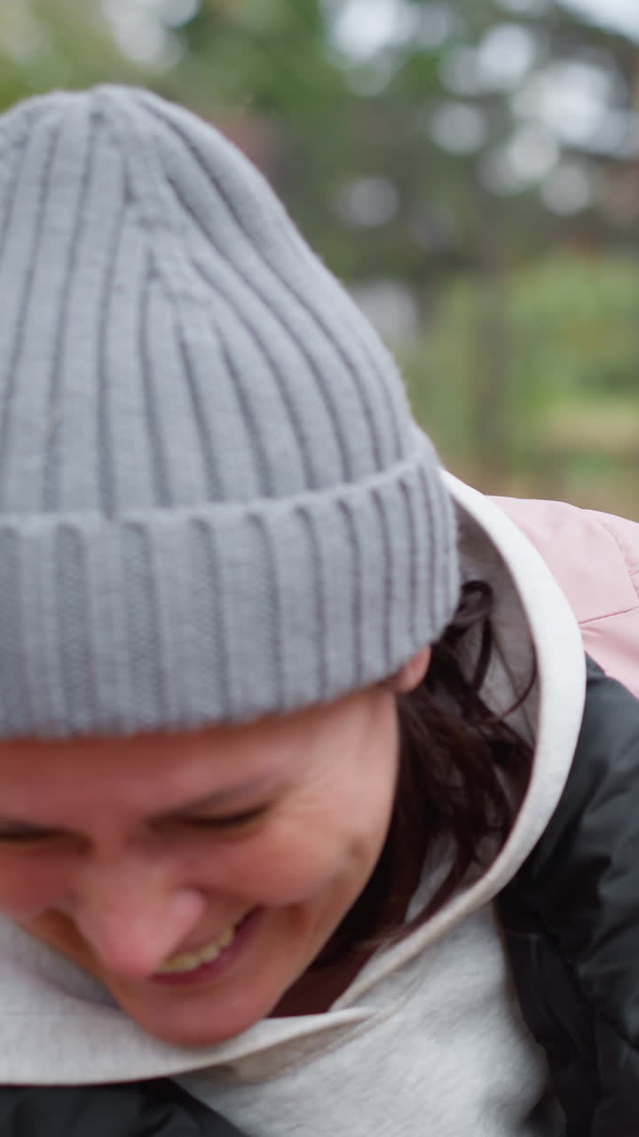 분홍색 재과 회색 모자를 입은 미소 짓는 여자가 밖에서 구부러져 건조한 잎자루가 놀랍게 그녀에 부어, 활기찬 가을 색상과 자연 풍경으로 둘러싸여 있습니다.