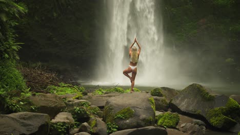 mujer rubia en equilibrio de pose de yoga de árbol con poderosa cascada nungnung