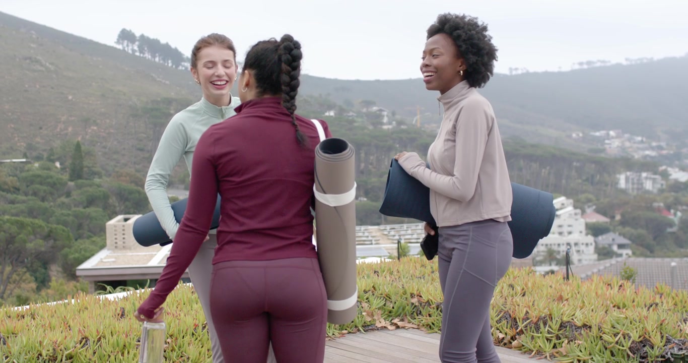 Diverse female friends walking on rooftop with mats and water bottle, preparing for yoga