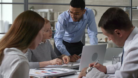 Businesswoman-In-Eyeglasses-Sitting-At-Meeting-Table,-Showing-Something-On-Laptop-And-Explaining-Document-To-Young-Employees-While-Working-Together-In-The-Office