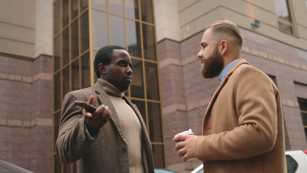 Bottom view of caucasian and african american businessman in elegant clothes talking in the street in autumn while one of them drinking coffee and the other holding a smartphone