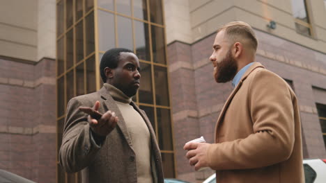 bottom view of caucasian and african american businessman in elegant clothes talking in the street in autumn while one of them drinking coffee and the other holding a smartphone