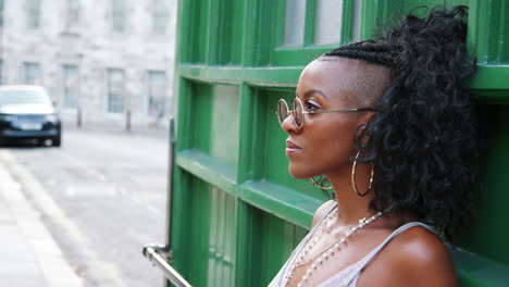 Moda-Joven-Mujer-Negra-Con-Gafas-De-Sol-Redondas-Y-Camisola-Apoyado-En-La-Pared-Verde-En-Una-Calle-De-La-Ciudad,-Vista-Lateral,-Cabeza-Y-Hombros