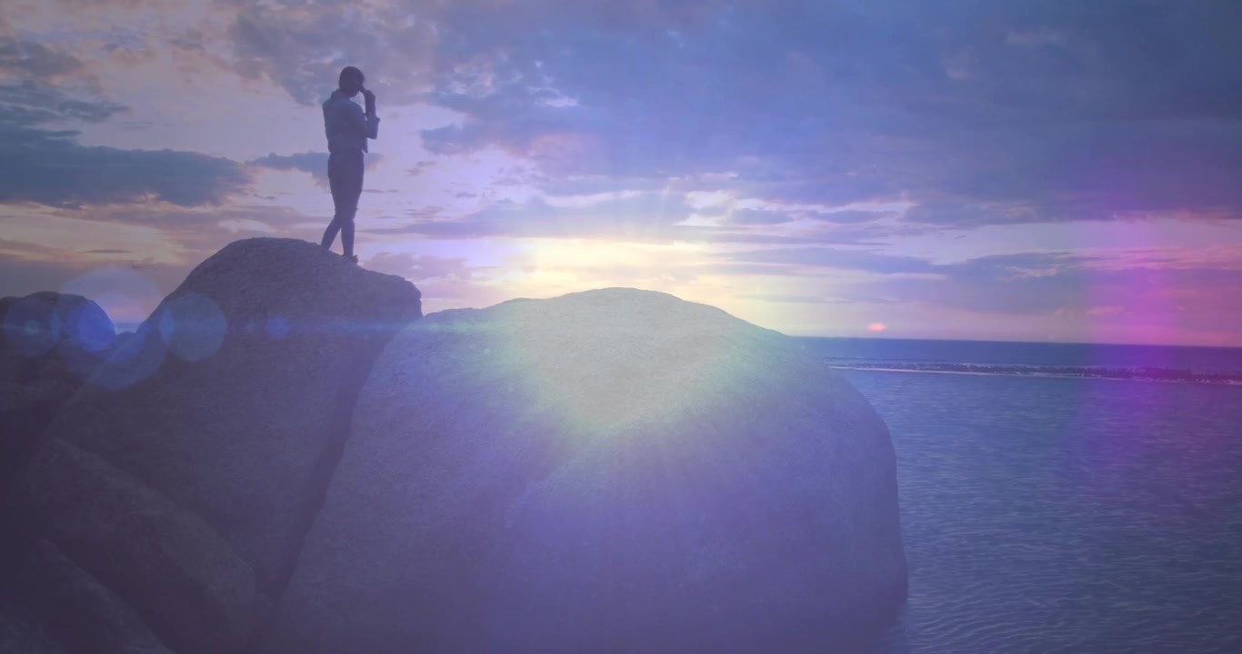 Viaggiatore che scala un masso al tramonto, sole che abbaglia, riparandosi gli occhi, tenendo lo smartphone e guardando il mare