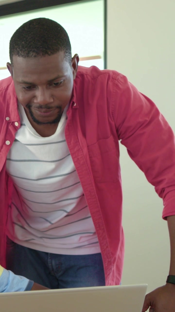 Vertical video: Leaning red-shirted father guiding son while typing on laptop at home, pencil shown
