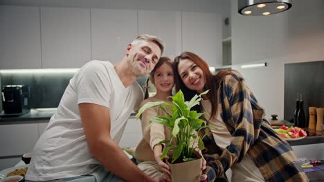 Portrait-of-a-happy-little-girl-with-her-parents-holding-a-houseplant-forward-and-rejoicing-after-her-dinner-in-a-modern-apartment-in-the-kitchen-in-the-evening