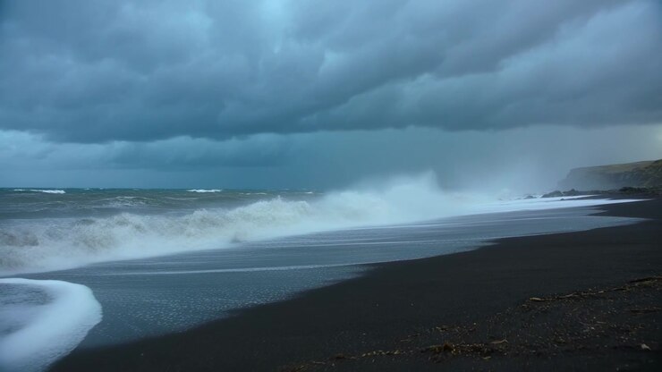 Houle montante approchant, la houle déferlant et se brisant sur une plage de sable noir, des embruns s'élevant près du cap.