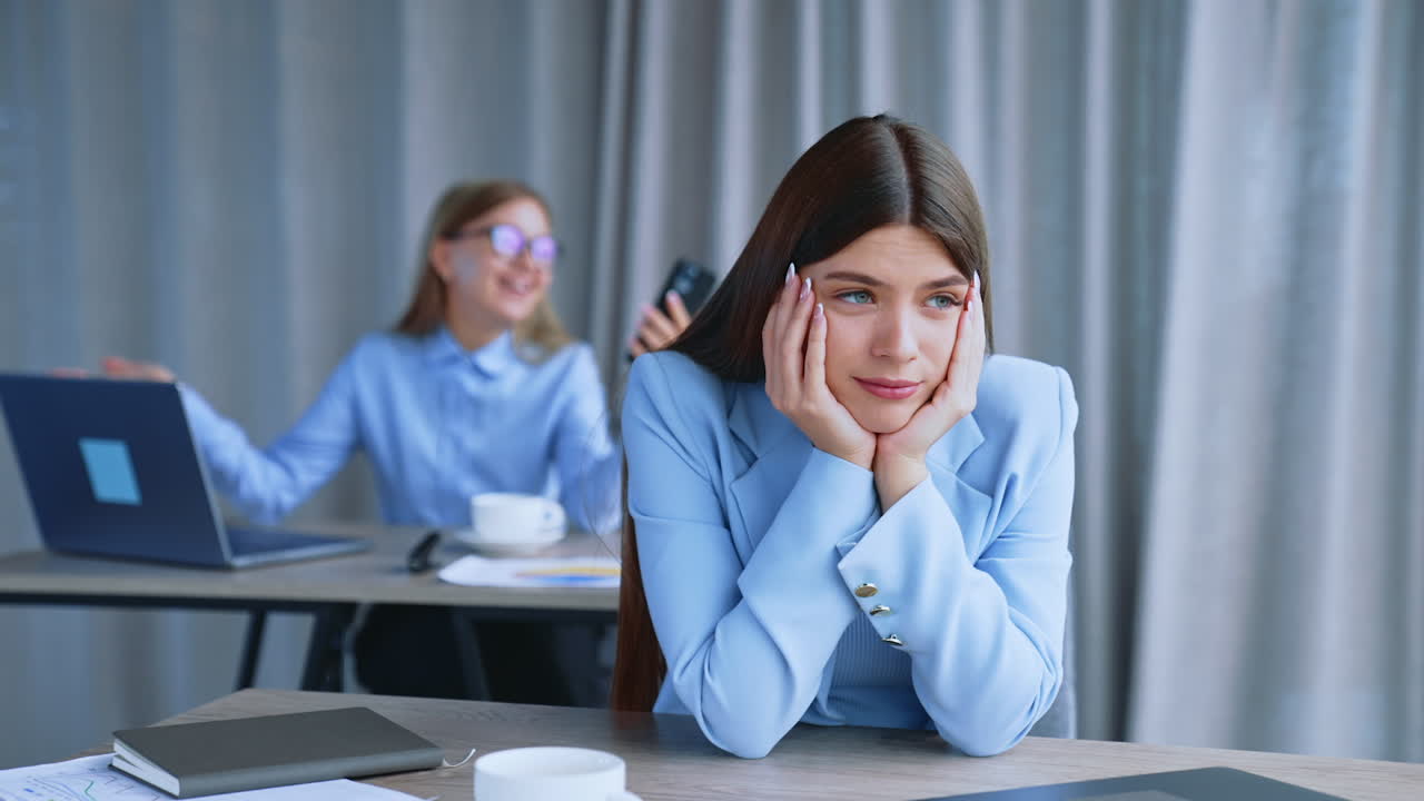 Beautiful long-haired brunette is sitting at desk in office. Female employee is bored having no work to do or doesn't like her job. Happy woman speaking on the phone happily at backdrop in blur.