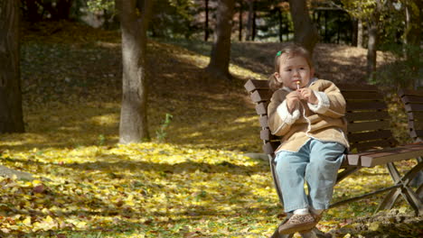 Niña-Comiendo-Caramelos-De-Piruleta-Sentada-En-Un-Banco-En-El-Parque-De-Otoño