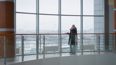 elegant lady in black jacket walking thoughtfully in modern mall, with glass walls and railing, cars visible through glass with blurred cityscape background, she holds a handbag, lost in thought