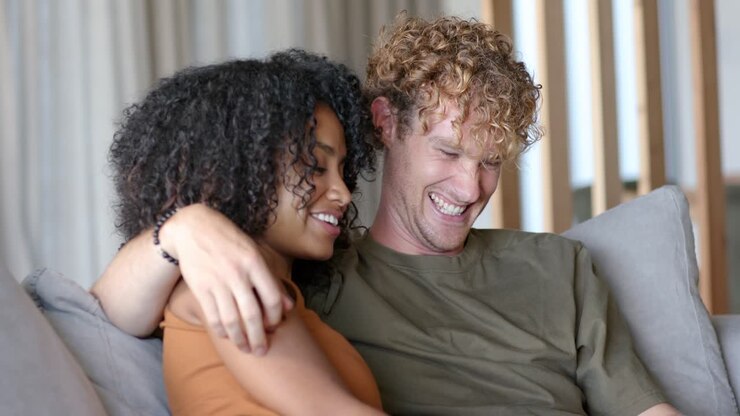Diverse couple after sitting cuddling on sofa, man draping arm, woman leaning, beaded bracelet