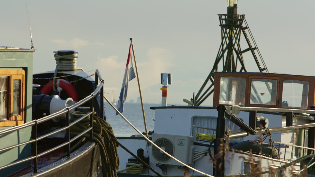 Barche da pesca ormeggiate in un porto con corde, dettagli del ponte, una bandiera olandese e un faro di navigazione, che rappresentano l'industria marittima, le infrastrutture costiere e la vita lavorativa del porto