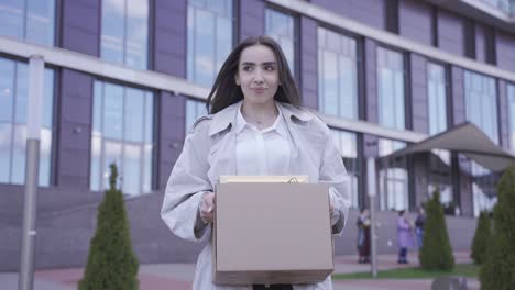 una mujer bonita camina frente a un edificio de oficinas. ella lleva sus cosas en una caja. ella ha dejado su trabajo. ella esta sonriendo.