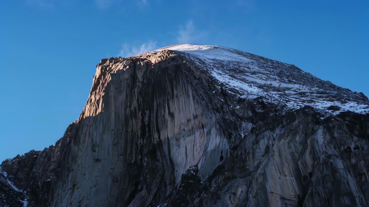 Affioramento di una cupola di granito dalla valle, sole basso che sposta la cresta e vento che solleva il pennacchio di neve dalla cima