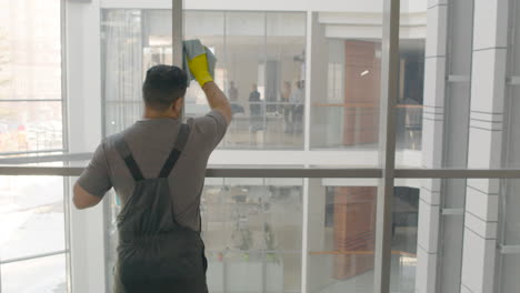 Rear View Of Arabic Cleaning Man Cleaning The Window Panes With A Rag And Glass Cleaner Inside An Office 1