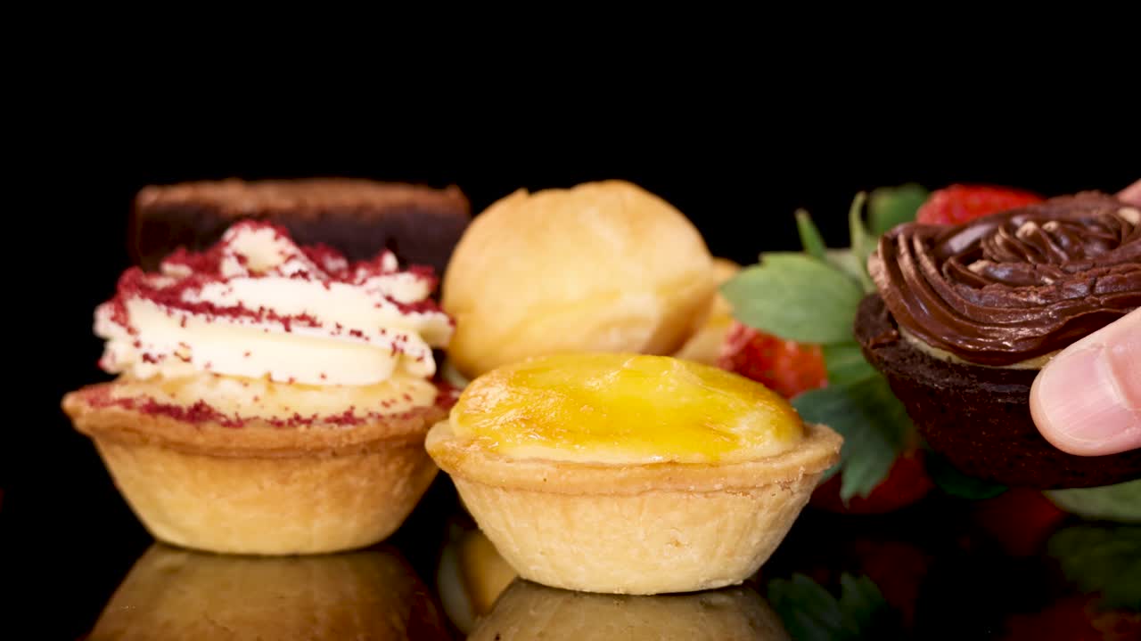 A hand picks up various mini pastries, including brownies, tarts, and cream puffs, from a glossy black surface with vibrant lighting and shallow depth of field