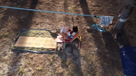 a young woman sits and reads a book close to her tent on a campsite in a wild pine forest