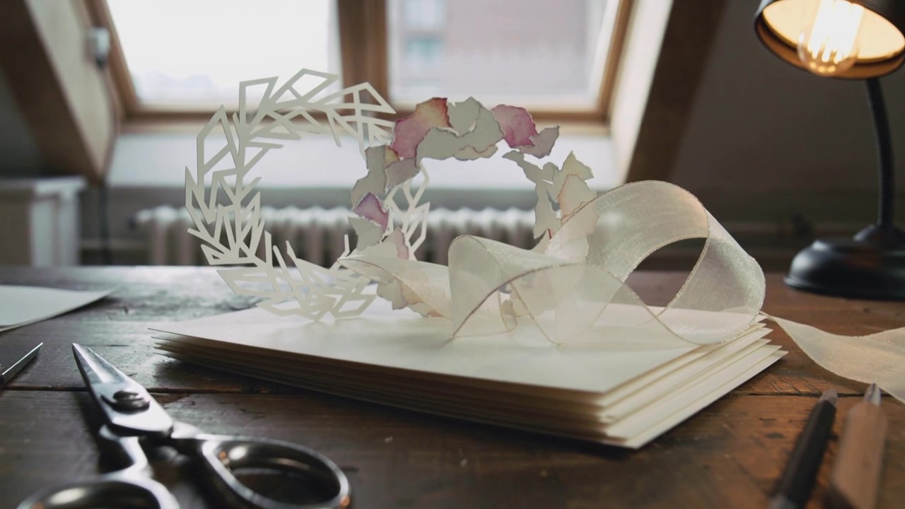 Pulling back camera revealing paper wreath on cream paper stack, breeze lifting ribbon, scissors