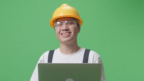 close up of asian man worker wearing goggles and safety helmet using a laptop and smiling while standing in the green screen background studio