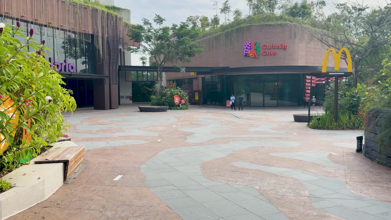 Un amplio plano panorámico revela la plaza de entrada de un zoológico moderno con vegetación y arquitectura contemporánea