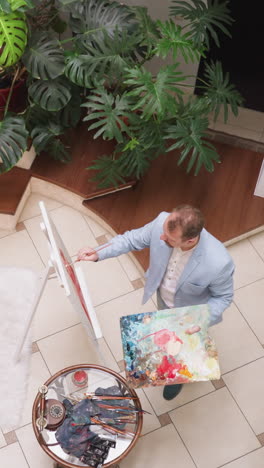 artist applies paint on canvas fixed on easel in plant gallery. professional painter holds palette drawing picture in home workshop. creative man improves talent