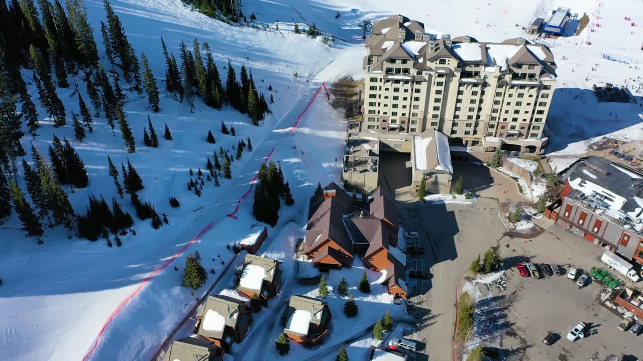 Aerial drone shot over Big Sky, Montana in winter, featuring rental condos near a ski resort, snow-covered peaks, pine forests, and stunning alpine scenery perfect for a cozy winter vacation retreat