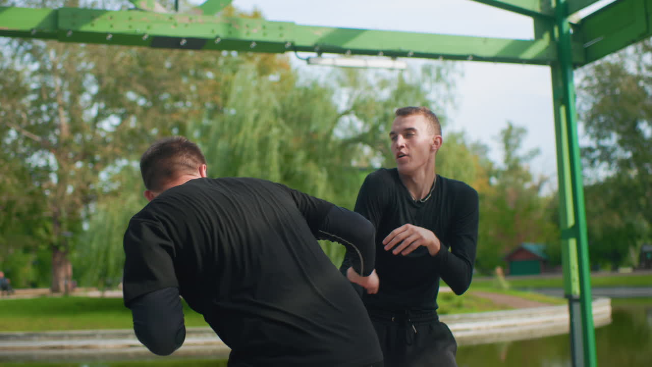 Caucasian athletes practicing boxing footwork drills in green pavilion near lake, focused coachlike partner guiding pivot and stance, repeated jab and guard adjustments, crisp rhythm and balance work