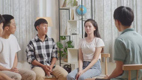 asian teen group sitting in chairs forming a circle discussing at home. everybody is listening to a girl talking about something