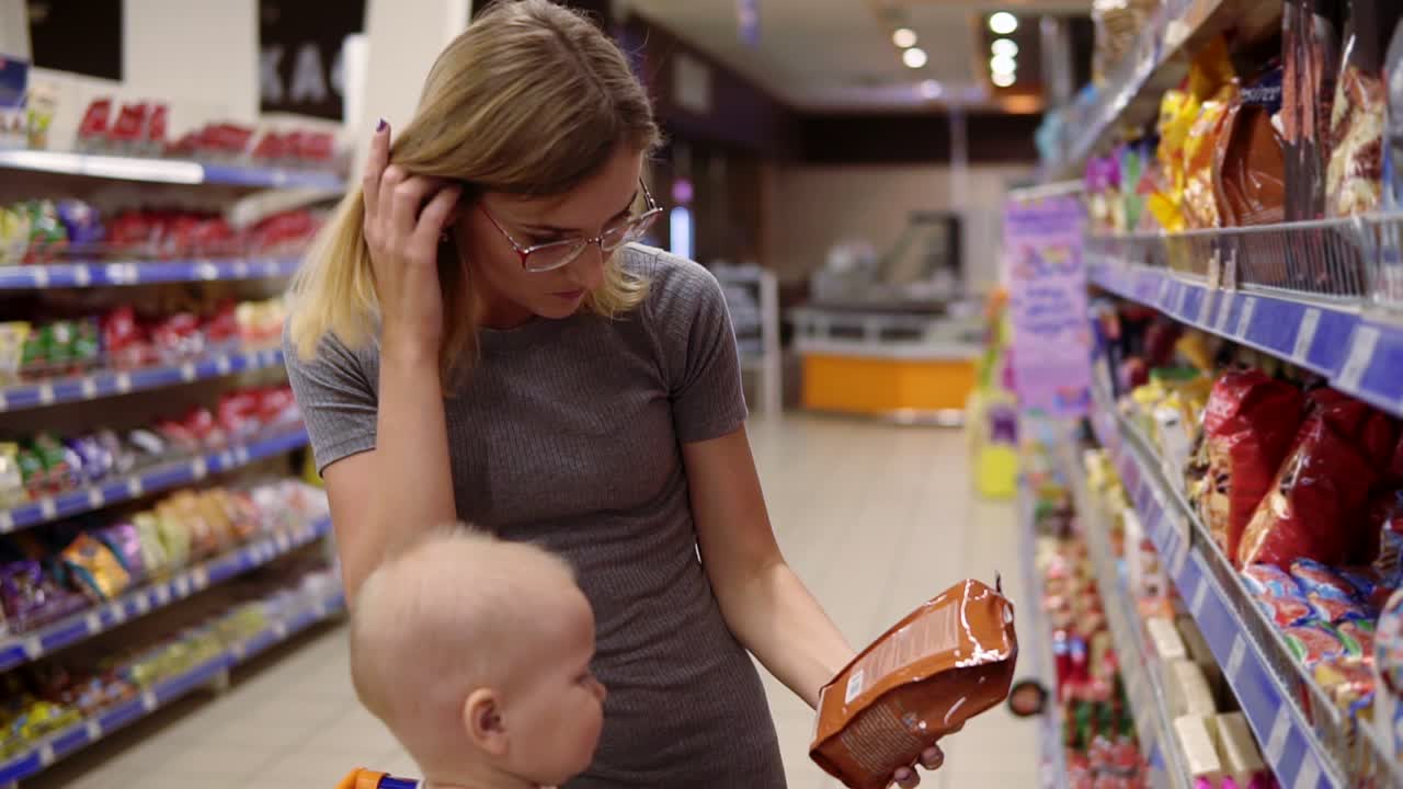 hermosa mujer está leyendo el contenido del producto en el paquete de galletas, mientras que su pequeño bebé está sentado en un carrito de comestibles en el