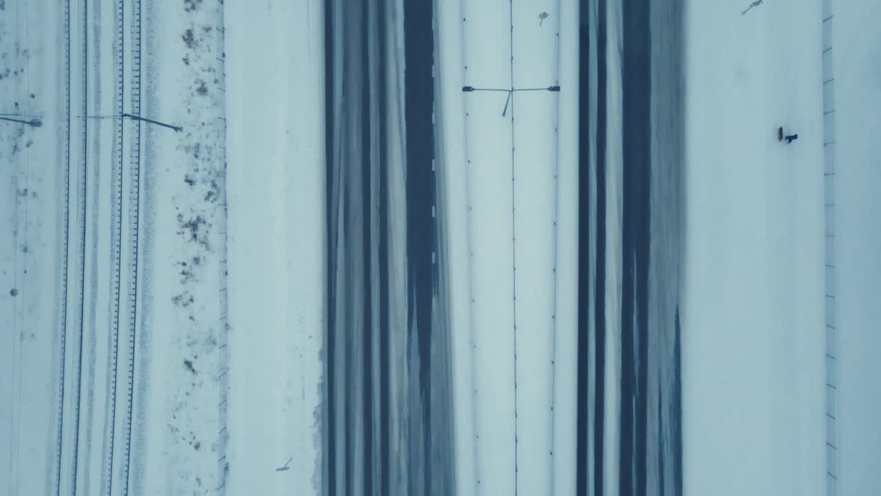 Drone flies above multi-lane highway with careful winter traffic, snowy surroundings, and rail tracks on a cloudy day