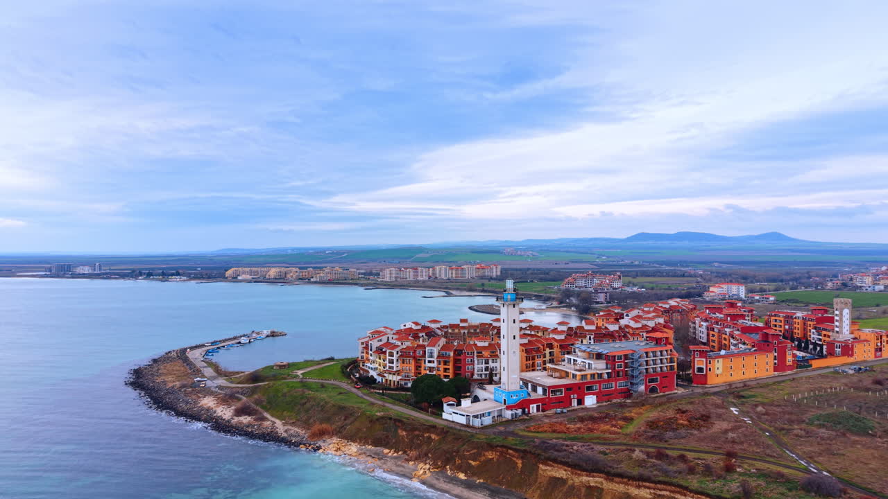 Vista aerea di una località balneare con faro ed edifici rossi. Veduta dall'alto di una cittadina costiera che mostra un faro bianco, architetture rosse e la costa sotto un cielo nuvoloso.