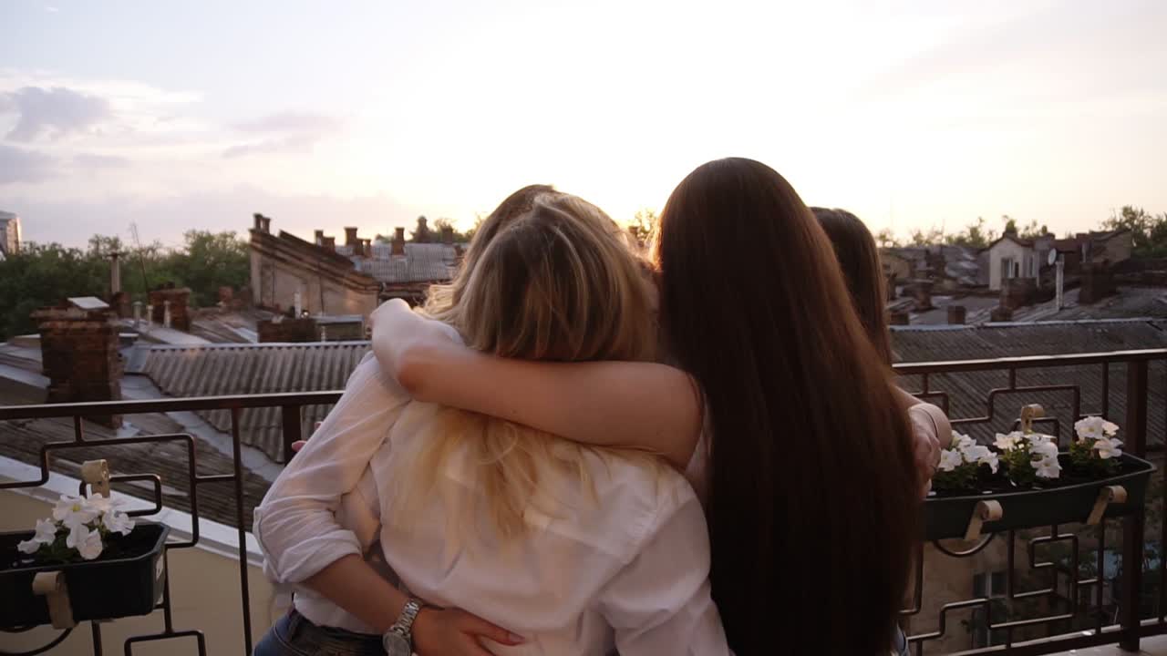 Happy, young girls embraced in circle, side view. Six attractive girls hugging, looking each other, standing outdoors on a terrace. Laughing. Town view on the background