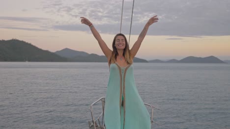 carefree woman standing and raising hands in the air on deck of boat with sunset sky in background