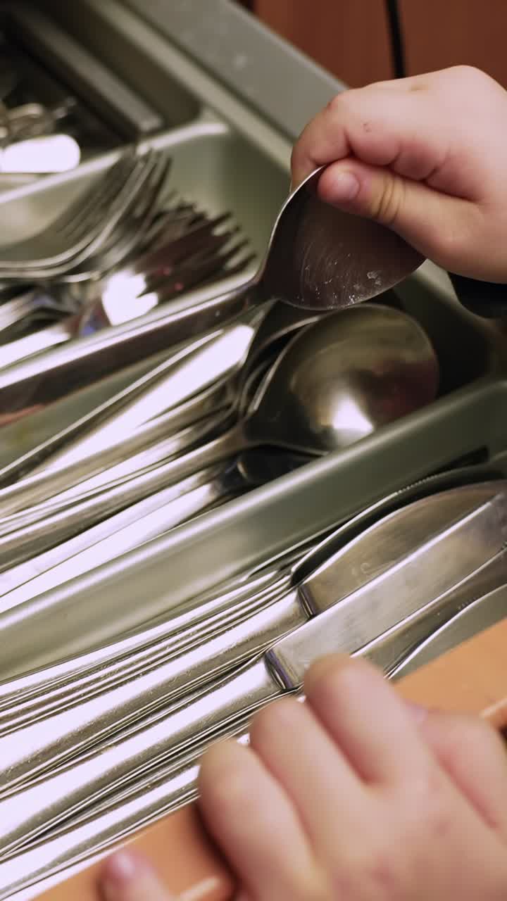 Vertical close-up of a child’s hand choosing a spoon from a cutlery tray in a kitchen drawer, showing everyday household organization and cleanup