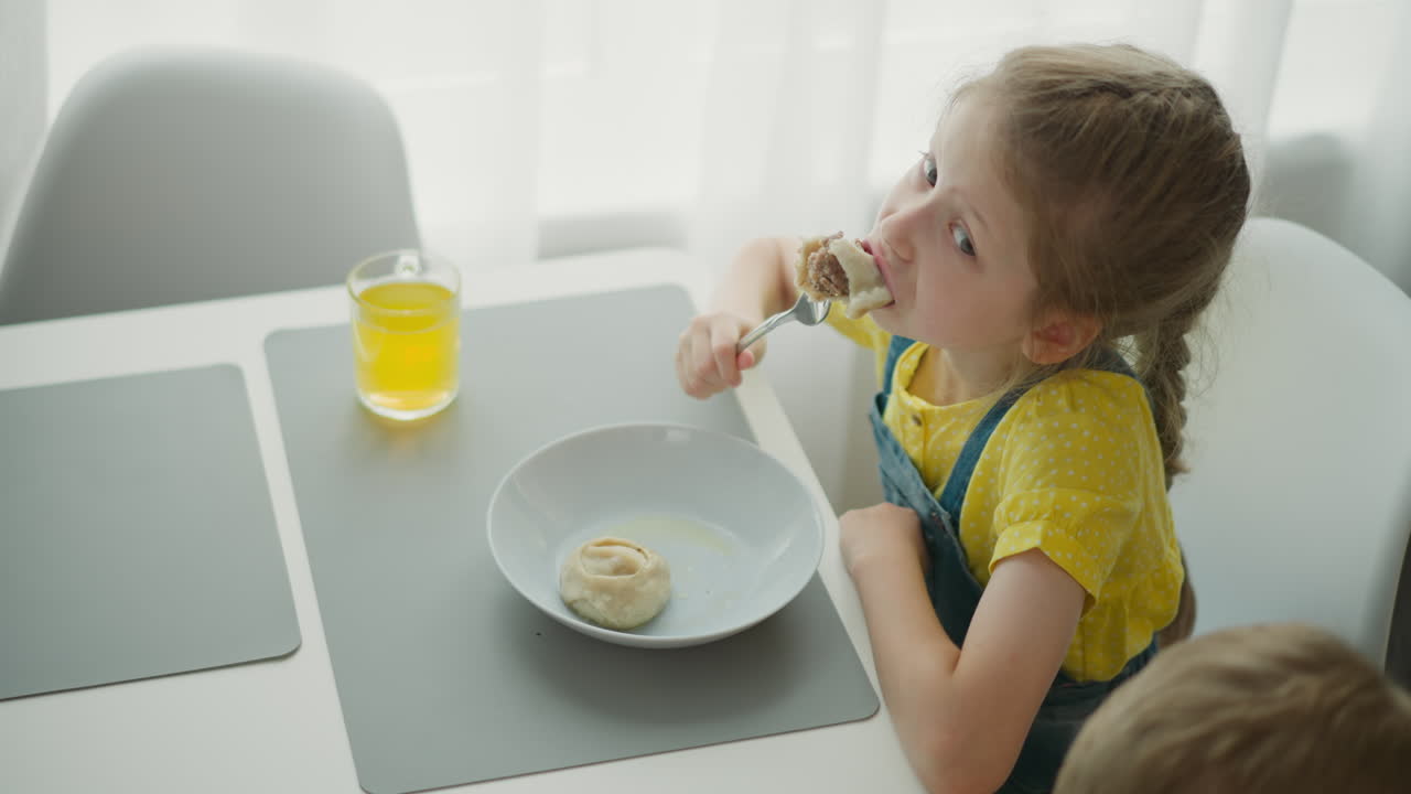 young girl wearing yellow top with blue overall sitting at dining table eating food from plate with fork, glass of yellow juice in front of her, partial head view of another person nearby