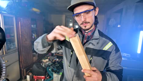 concentrated young bearded joiner in goggles and a cap checks the work piece