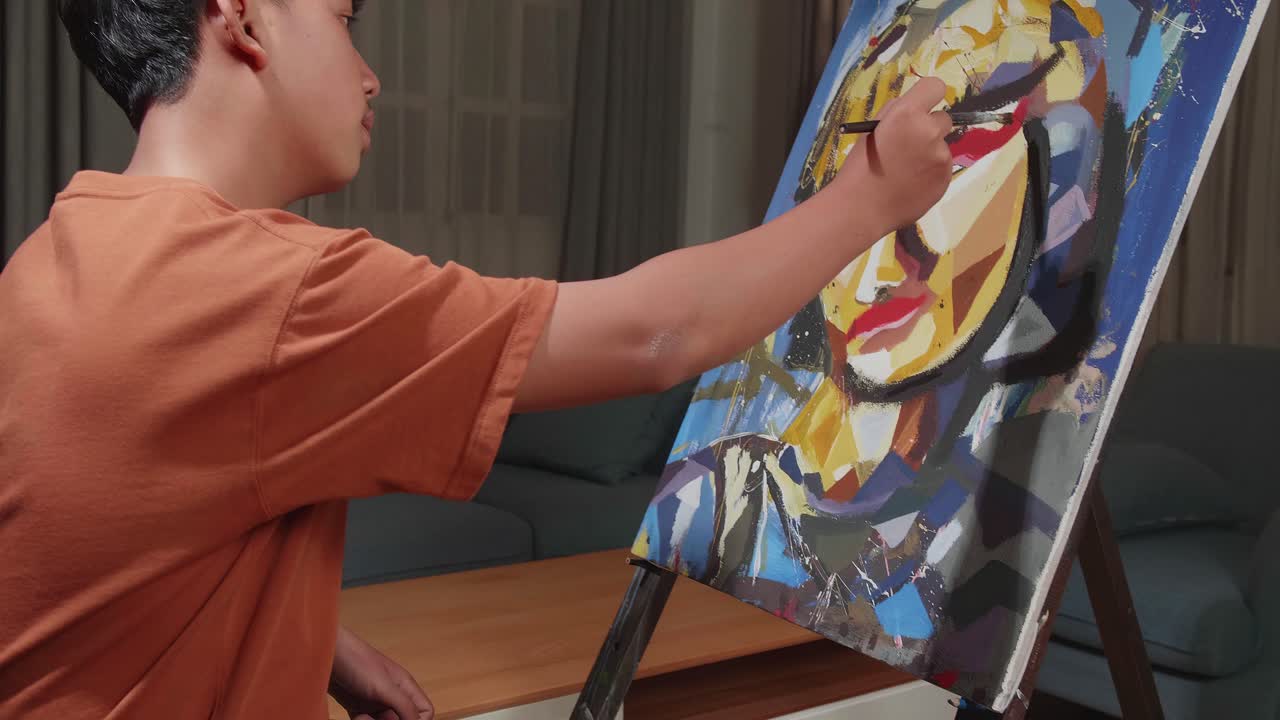 Asian Artist Boy In Wheelchair Holding Paintbrush And Mixing It With Colour Before Painting A Girl'S Face On The Canvas