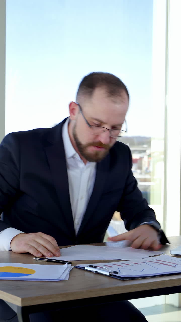 Nervous businessman in office. Irritated man entrepreneur sitting at table and tearing papers throwing them to the sides on the background of window. Vertical video