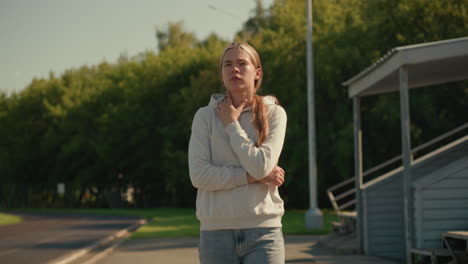 girl in casual hoodie, walking outdoors on sunny day near stadium, touching her neck in thoughtful manner, background features electric poles, lush green trees, and an empty stadium