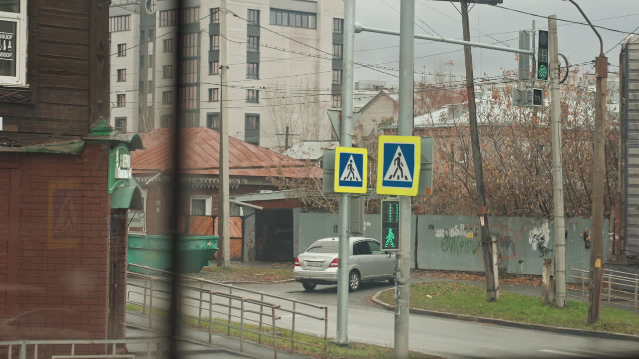 City street crosswalk seen from window, creator filming white pedestrian under overcast sky, cars passing along wet road, apartment blocks and traffic signals in background, muted color palette