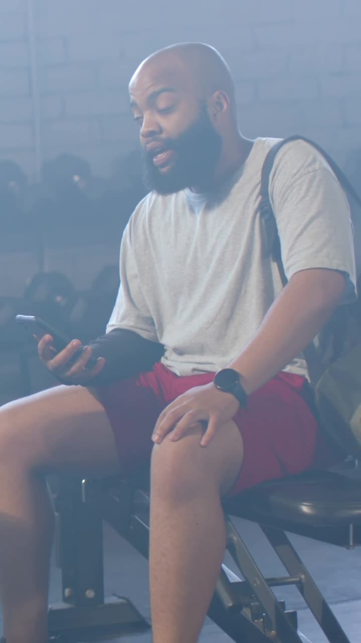 Video vertikal: Seorang pria mengenakan kaus abu-abu dan celana pendek merah sedang mengetuk dan memeriksa ponsel pintar di bangku gym, sambil tersenyum.