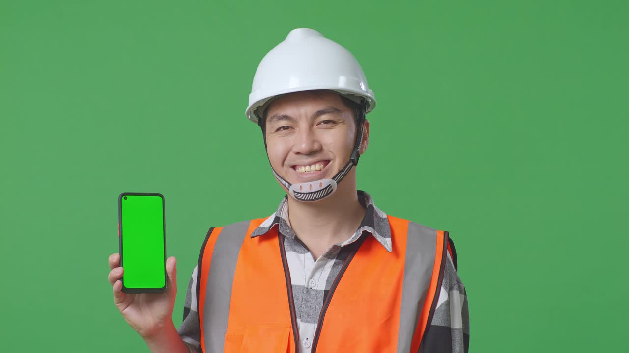 Close Up Of Asian Male Engineer With Safety Helmet Smiling And Showing Green Screen Smartphone To The Camera While Standing In The Green Screen Background Studio