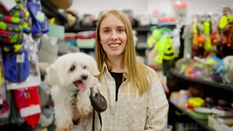 Portrait-of-a-happy-blonde-girl-in-a-checkered-light-shirt-with-her-white-cheerful-dog-in-a-pet-store.-Portrait-of-a-happy-girl-who-came-to-the-pet-store-with-her-dog