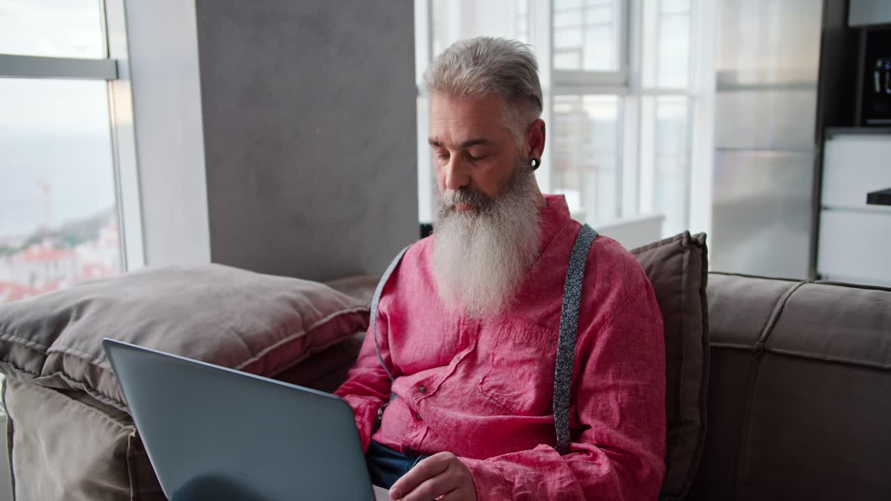 A confident and happy man with gray hair and a lush beard of advanced age in a pink shirt sits on a brown sofa and types on his gray laptop in a modern apartment