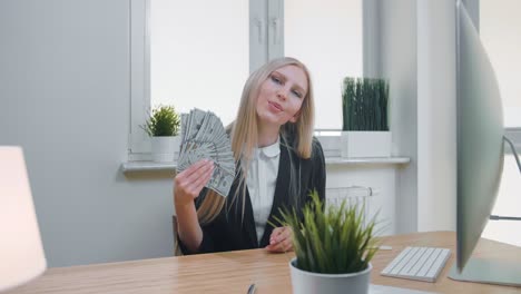 relaxed woman with money in office. elegant young blond female in business suit sitting at desk with computer and plant and holding in hand fan of cash confidently looking at camera.