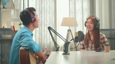 young asian woman in headphones singing into microphone while man playing the guitar during online livestream in podcast studio