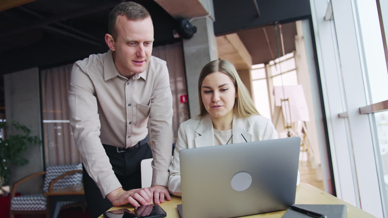 A couple of colleagues discuss the issue looking at laptop. Man bent to computer on the desk next to a sitting lady. Positive pleasant talk at an office.