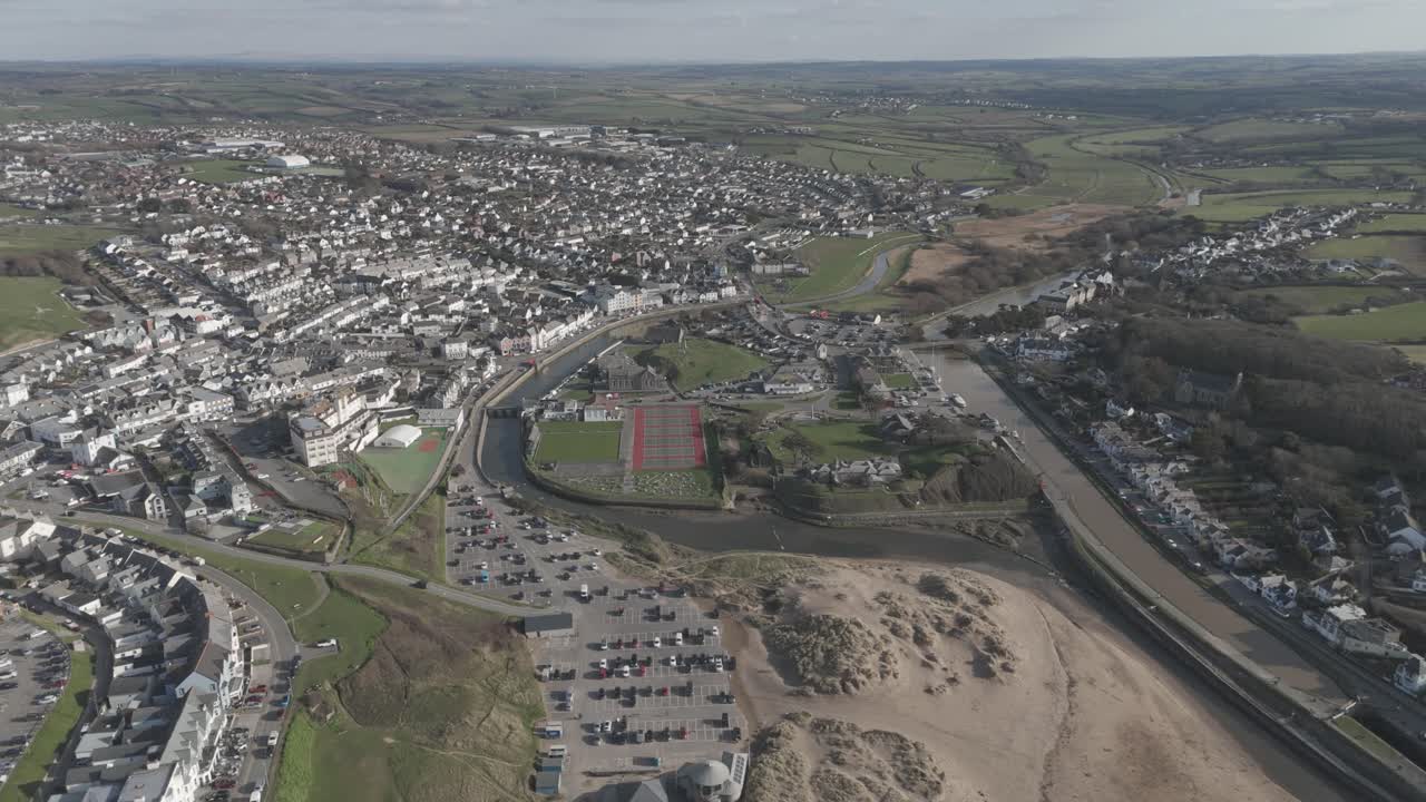 Aerial sweep over Bude town unfolding from tidal river and pastel rooftops to dunes and Atlantic surf, capturing coastal community, recreation and natural scenery