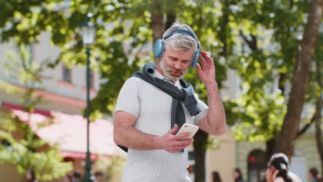 Happy-mature-man-in-wireless-headphones-choosing,-listening-music-in-smartphone-application-outdoors