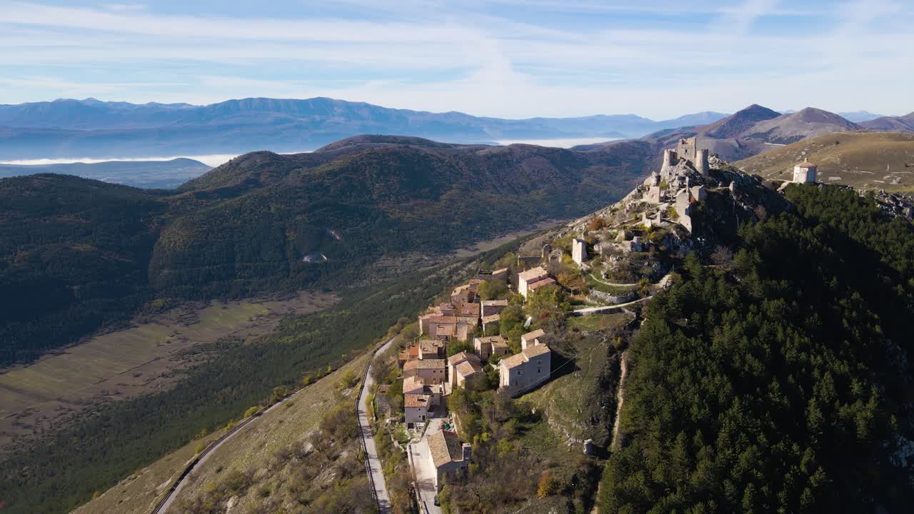 Splendida vista aerea del borgo medievale di Rocca Calascio in cima a una collina che scende lungo un crinale roccioso con rovine della fortezza sopra, nebbia nella valle sottostante e la drammatica catena montuosa del Gran Sasso oltre